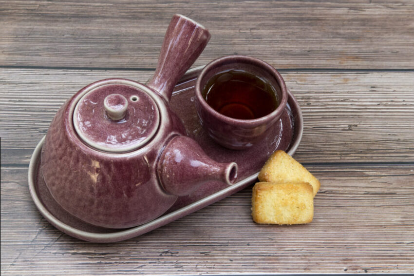 A beautiful, set of purple ceramic Chinese style tea pot and tea cup with black tea and pieces of sugar biscuits on brown wooden background.

