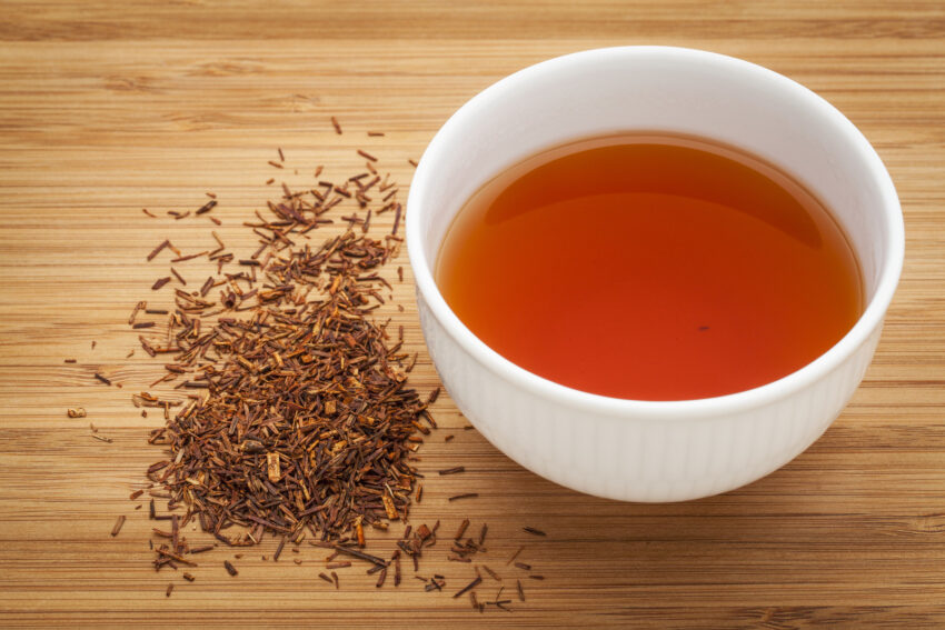 Rooibos red tea - a white cup of a drink and loose leaves on bamboo wood background, tea made from the South African red bush, naturally caffeine free